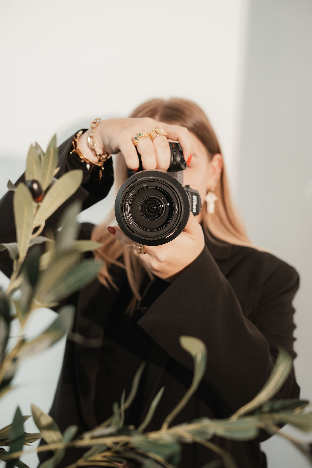 Lara Haymoz Une femme debout tient un appareil photo entre ses mains, prête à capturer l’instant. Derrière elle, un fond à rayures bleues et blanches apporte une touche graphique et dynamique.